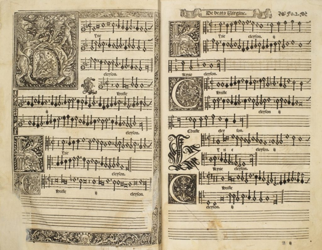 First page of Morales's Missa de Beate Virgine, depicting an illuminated K with a vignette of the Virgin and child in the center. The next verse features a smaller illuminated K depicting a cherub playing a harp. On the opposite page, the same smaller K design is repeated, followed by verses that feature small illuminated letter C's, each surrounded by a decorative floral design.